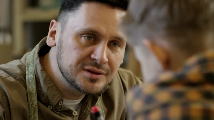 Tilt down of professional male carpenter drawing pattern on wood plank and talking to unrecognizable little son in workshop