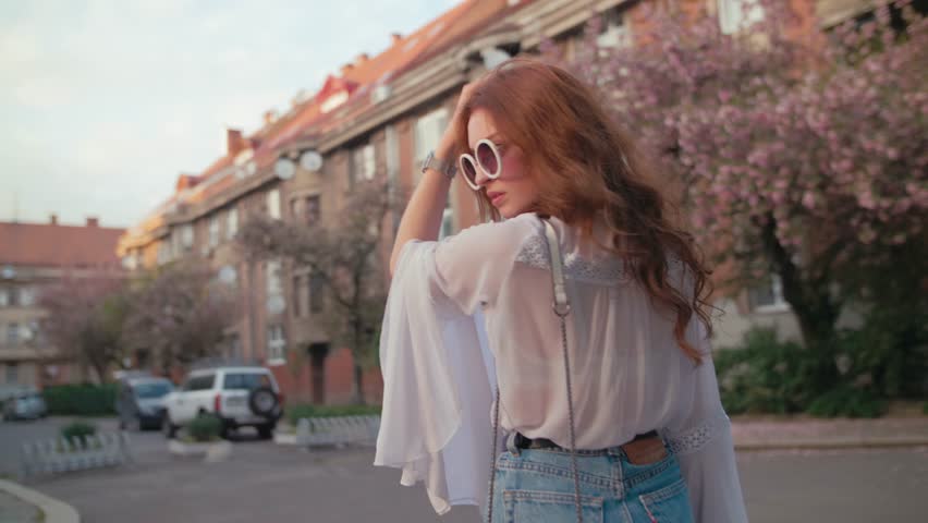 Young beautiful fashionable woman wearing stylish round sunglasses, white chiffon blouse, wrist watch, with small bag posing in street of city