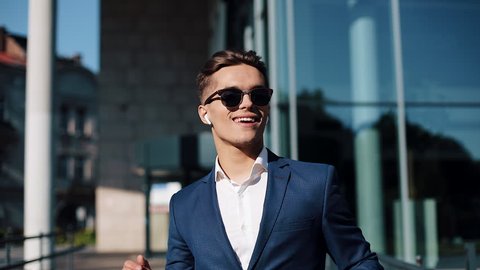 Young businessman with sunglasses listening to the music on his smartphone outdoors and dancing. He walking near office building. Communication, audiobooks, music, business people. Shot on Red Epic