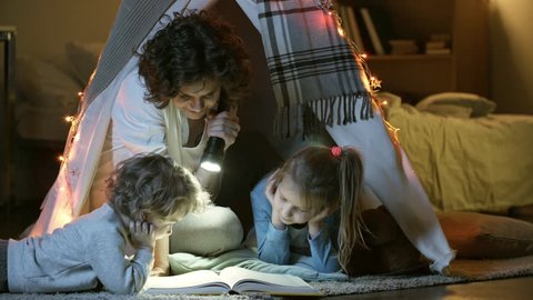Two little children of primary school age looking at book of fairy tales when sitting in teepee play tent with loving mother