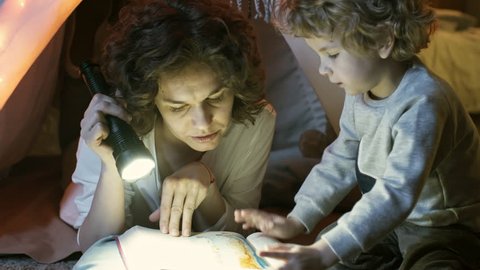 Close up shot of cute little boy pointing at something in fairy tale book and talking to caring mother when reading together using flashlight