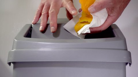 Slow motion close shot of a man’s hands putting orange peel from a piece of kitchen towel into a plastic swing bin / trash can.
