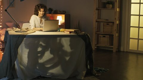 Tracking shot of smiling young woman working from home with laptop computer and looking after two little children playing under desk