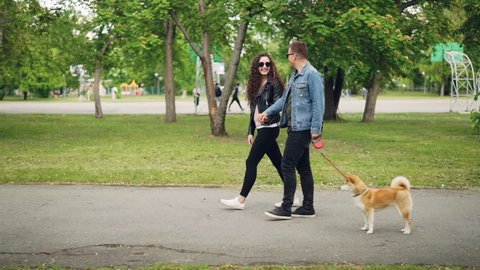 Slow motion side view of attractive young man dog owner walking his pet and chatting to his girlfriend going hand in hand with her. Active lifestyle and animals concept.