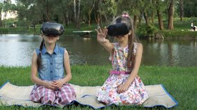 Children sit in the Park on the grass by the river in virtual reality glasses and play virtual games. They wave their hands and laugh. 4K, 29.97 fps.