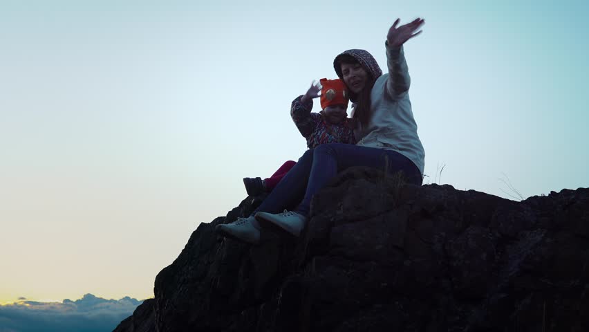 Family travel in the mountains, mother and daughter are sitting on a rock against the blue sky at sunset. They smile and wave their hands.