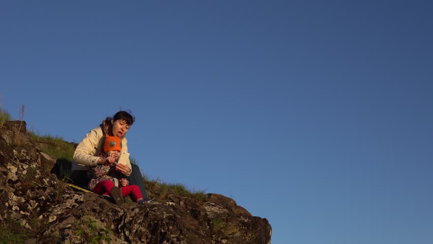 Family travel in the mountains, mother and daughter are sitting on a rock against the blue sky. A young woman and a little girl are recording video using a smartphone, they are waving their hands.