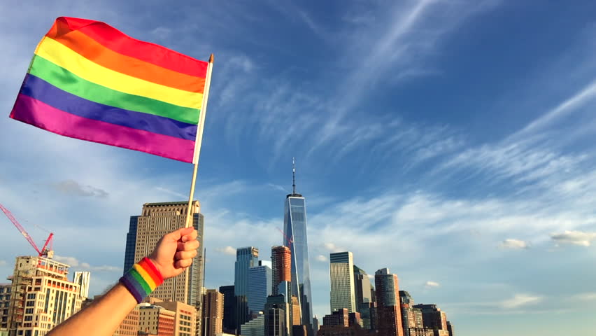 Hand wearing gay pride wrist band holding rainbow flag up in front of the city skyline at sunset Royalty-Free Stock Footage #1012822793