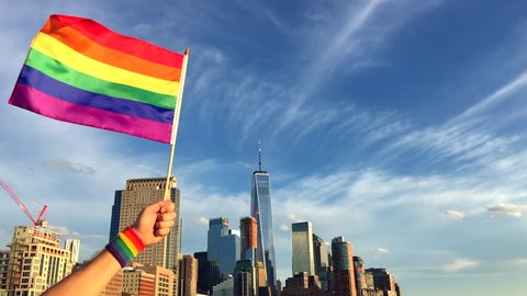 Hand wearing gay pride wrist band holding rainbow flag up in front of the city skyline at sunset