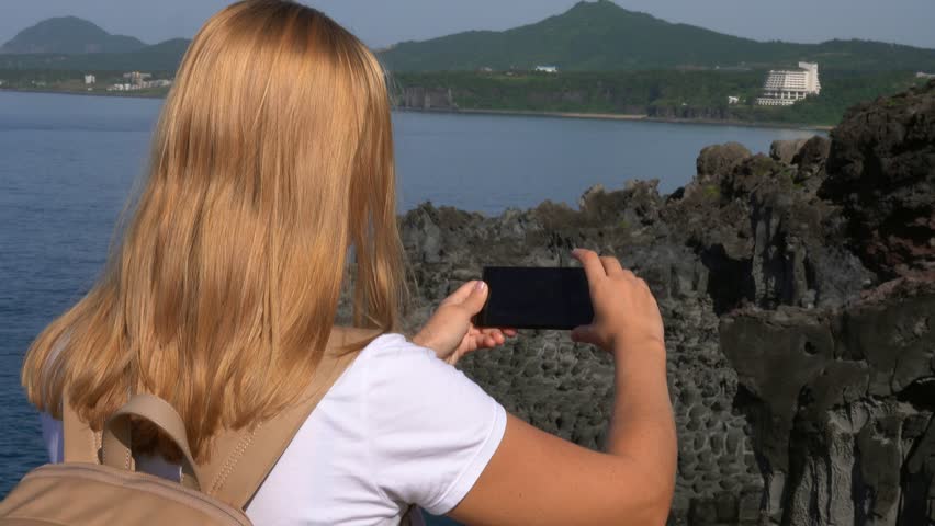 Caucasian Female Tourist Taking Photo of Daepo Jusangjeolli Cliff on Jeju Island (South Korea) with Mobile Phone. Active Tourism in Asia