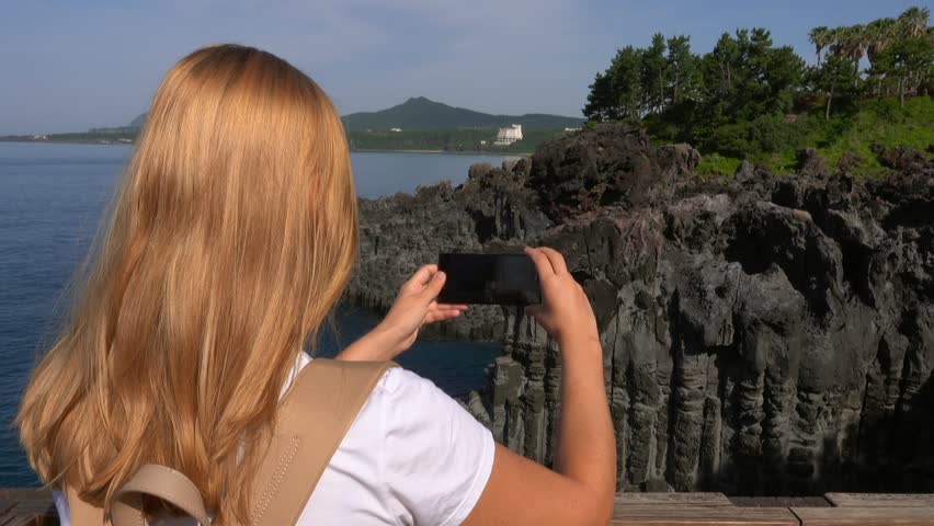 Caucasian Female Tourist Taking Photo of Daepo Jusangjeolli Cliff on Jeju Island (South Korea) with Mobile Phone. Active Tourism in Asia