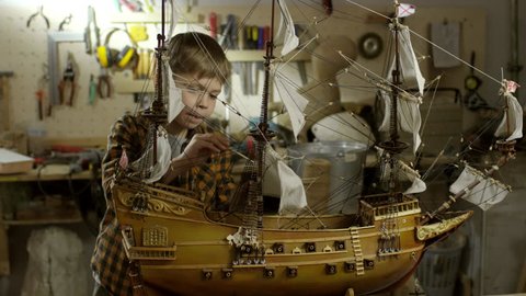 PAN of concentrated little boy applying paint coat on ship model standing on workbench in garage