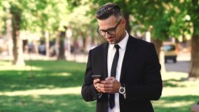 Close up view of happy businessman dressed in shirt using smartphone while walks in park