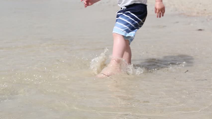 Little child boy run barefoot on the white sand beach near blue calm sea, close up on legs and feet.