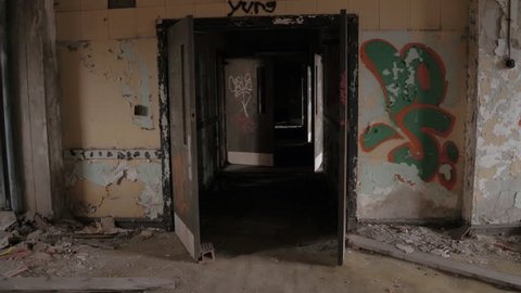 Detroit, United States, 18th October 2016, FPV, CLOSE UP: Walking along dark narrow hallway in abandoned decaying psychiatric hospital. Moving past the weathered wooden doors and broken walls.
