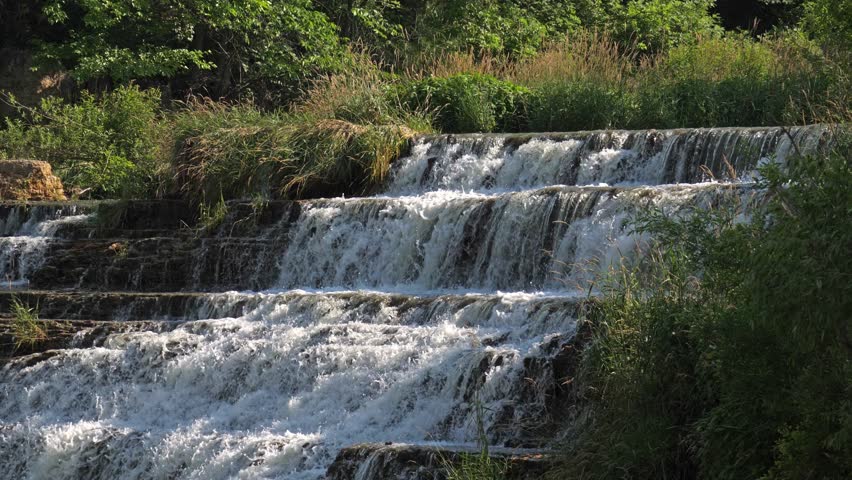 Slow motion view panning down the rocky stepped cliff of the beautiful Thunder Bay Waterfall as water pours down into the river below in Galena Illinois.