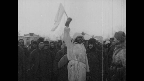 1930s: Man holds white flag. Large crowd of surrendered soldiers walks down street. Spectators.