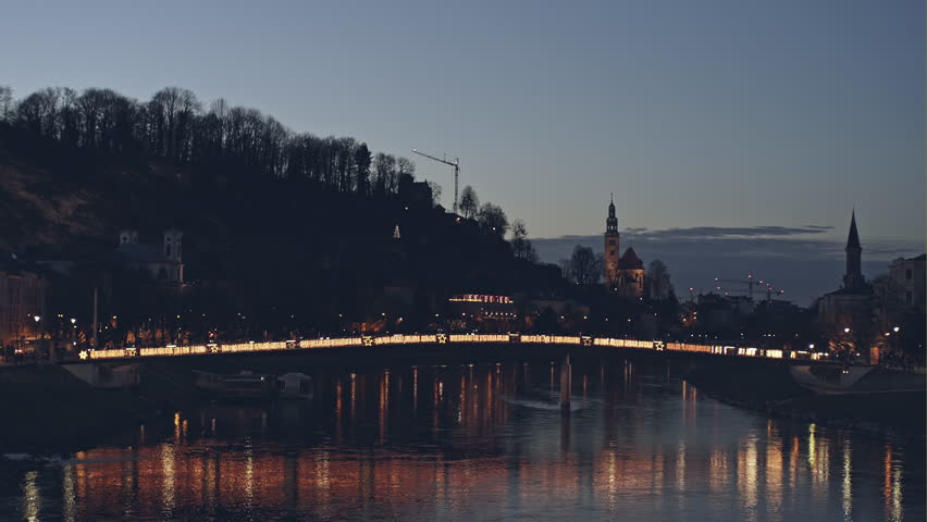 Magnificent view of bright illuminated bridge across small river at night with old stunning buildings of Salzburg and trees on background in Austria
