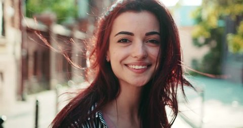 Close up view of happy young woman with red hair and big brown eyes, standing in the old city quarter, strong wind plays with her hair, she cheerfully smiles straight to camera. Female portrait