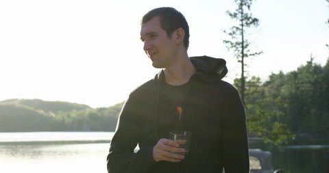 Young 20s man holding alcholic drink at cottage chats with friends off camera - wide shot