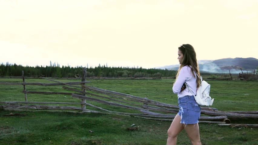 A 20s attractive brunette girl with a white backpack walks by an old fence outdoors 4k.