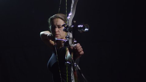 Studio shot of determined female archer with bow shooting arrow against black background