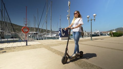 Full shot - Female riding electric scooter through marina. Modern transportation gadget and popular futuristic device among young people