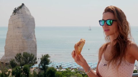 Woman eating croissant in front of sea. Beach of Pizzomunno famous white rock, Puglia, Italy.