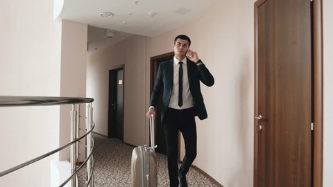 Happy young businessman at hotel lobby using smartphone, walking with luggage suitcase to airport terminal, or modern office