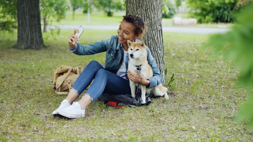Pretty young girl blogger is taking selfie with purebred dog outdoors in city park cuddling and fondling beautiful animal. Modern technology, loving animals and nature concept. | Shutterstock HD Video #1012911764