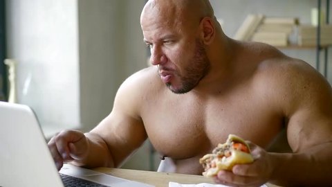 male athlete with big muscles sitting at the table working on laptop and eating triple Burger with beef Patty