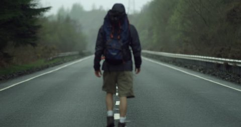 4K Young Traveller with Backpack walking on a Highway in the Scottish Glen Coe Mountain Highlands. Man walking on a Concrete Road within Forest and Mountains in Rural Scotland.