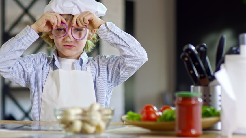 Portrait of playful little boy in apron and chef hat holding circles of red onion over eyes, smiling and looking at camera
