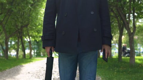 A young man in a black coat walks along the alley of flowering apple trees in the park and holds an umbrella and a mobile phone in his hands.