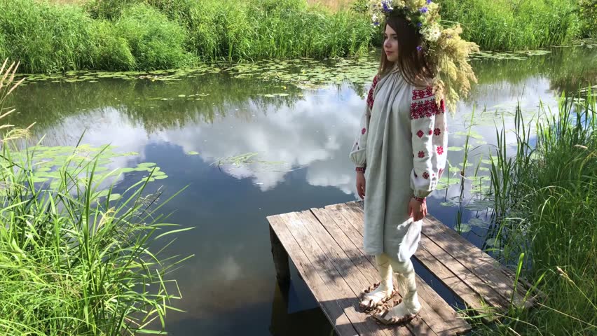 the girl sits on a wooden masonry on the river. A girl with a wreath on her head near the river. Kupalle day.