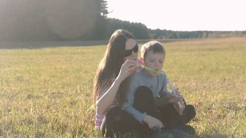 Mom and son blowing a soap bubbles sitting on the grass, Family weekend in park.