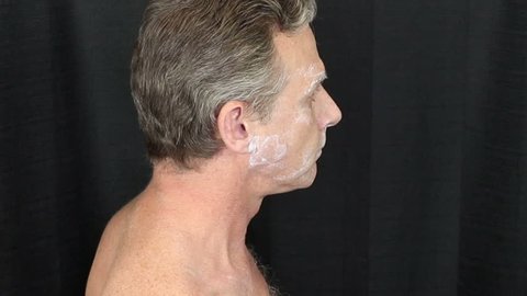 Man with thin white bentonite clay face mask turning back and forth in front of a black curtain. Shirtless male turning left and right while wearing a white clay bentonite face mask beauty treatment.