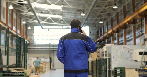 Engineer in in blue uniform is Walking Through Factory and talking on the mobile phone. Back View