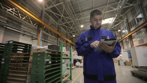 A man is working in a factory with a digital tablet, he is dressed in a blue uniform. factory worker.