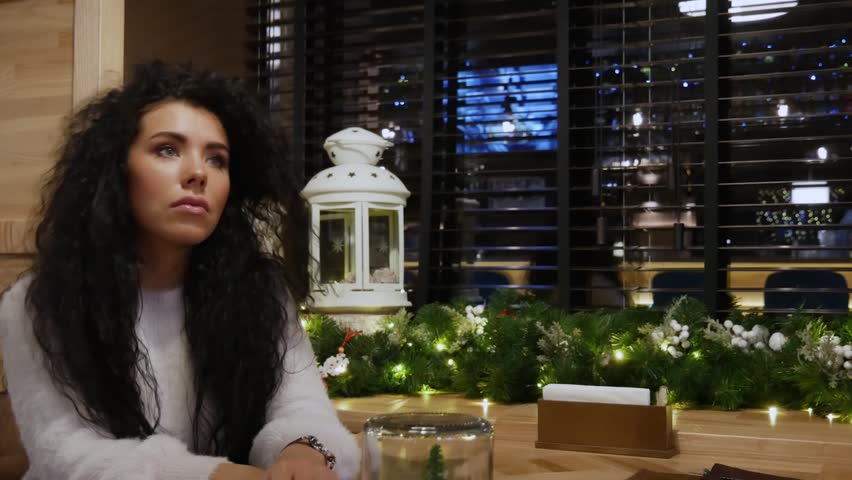 Beautiful curly-haired girl is waiting in the restaurant and smiling. Cute brunette in a white sweater is enjoying herself in a winter cafe