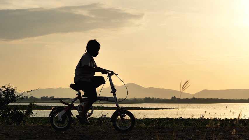 Silhouette of boy of 7 years old having fun riding his bicycle in time sunset. Slow motion