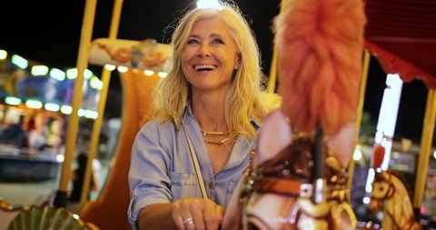Happy fashionable senior woman on summer vacations having fun on funfair merry-go-round ride at night