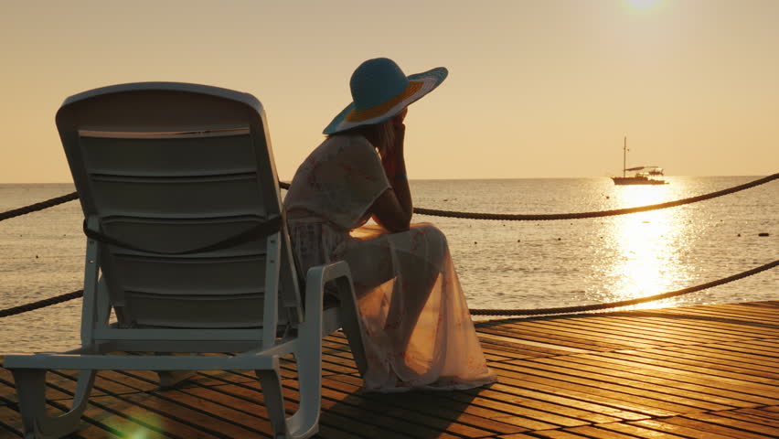 A woman looks at the sunrise over the sea. Sits on a sunbed, in the distance a fishing boat is visible. Scenic dawn landscape