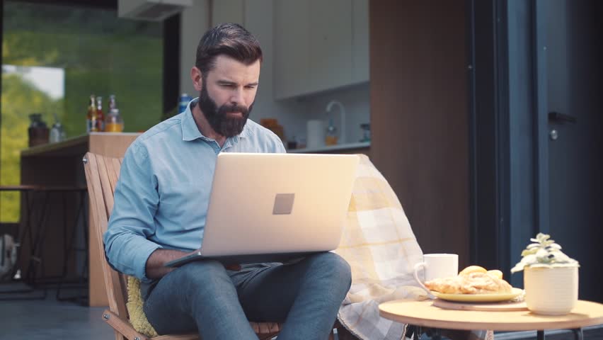 Attractive focused brown-haired man with beard wearing shirt and trousers looking at screen of laptop. Serious businessman working at home, holding cup in hand. Outdoors. Royalty-Free Stock Footage #1013039051