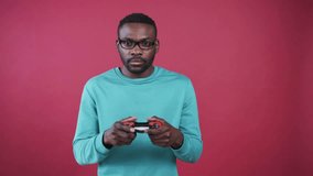 Cute focused African student holding gamepad in hands, playing video game. Young handsome man in black spectacles looking attentively at screen. Leisure. Pink wall.