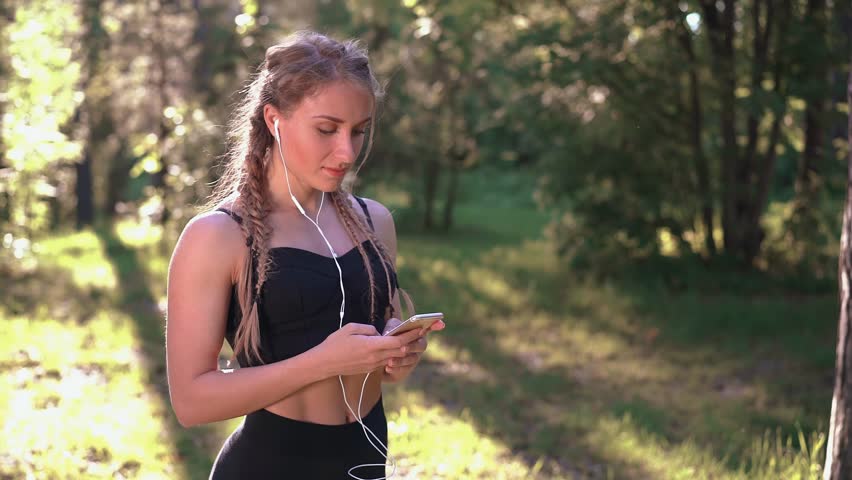 Attractive young woman in sportswear choosing music before her workout in the park on a sunny summer day. Smartphone and headphones. Left to right pan slow motion medium shot