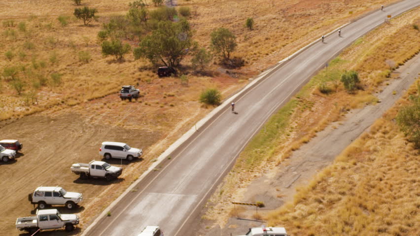 A birds eye view shot of a long road and trees with a person cycling in slow motion. Camera slowly moves forward.
