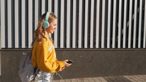 Portrait of young cute attractive young girl in urban background listening to music with headphones. Woman wearing yellow blouse and silver skirt.