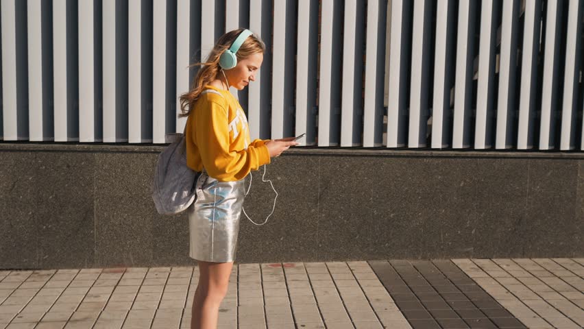 Portrait of young attractive teenager woman in urban background listening to music with headphones. Woman wearing yellow blouse and silver skirt. | Shutterstock HD Video #1013067575