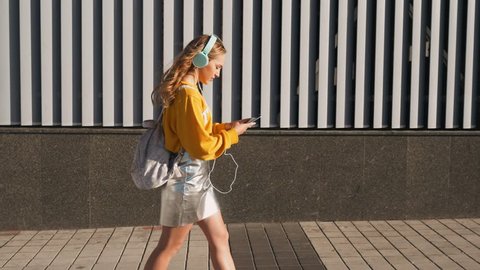 Portrait of young attractive teenager woman in urban background listening to music with headphones. Woman wearing yellow blouse and silver skirt.